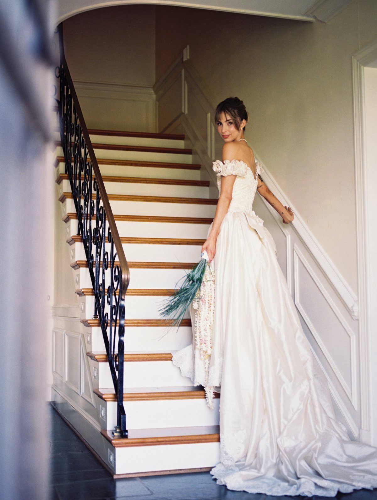 1989 Wedding Dresses: 8 Details That Defined the Look 6 A woman in a white off-the-shoulder wedding dress standing on a staircase holding a bouquet.
