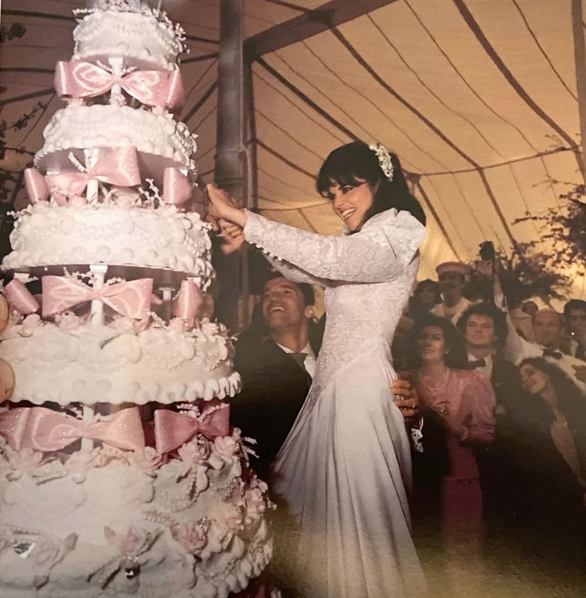 1989 Wedding Dresses: 8 Details That Defined the Look 5 A bride in a lace wedding dress cuts a large tiered cake decorated with pink bows.