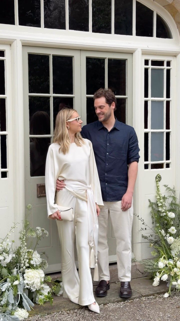 After Wedding Dress Ideas: 5 Looks Beyond the Ceremony 4 A woman in a cream silk jumpsuit and a man standing by white doors.