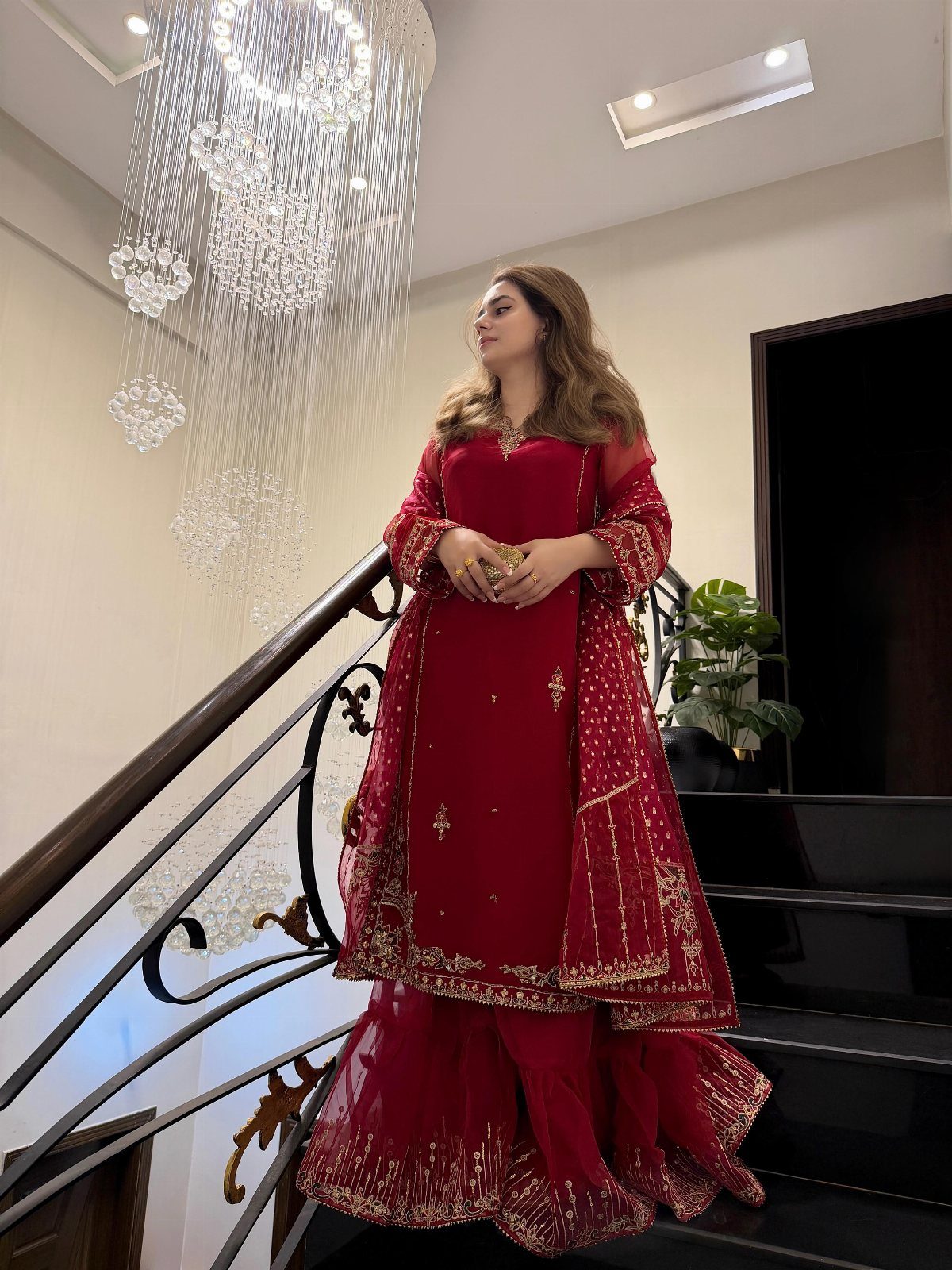 After Wedding Dress Ideas: 5 Looks Beyond the Ceremony 5 A woman in a red embellished traditional South Asian outfit posing on a staircase near a chandelier.