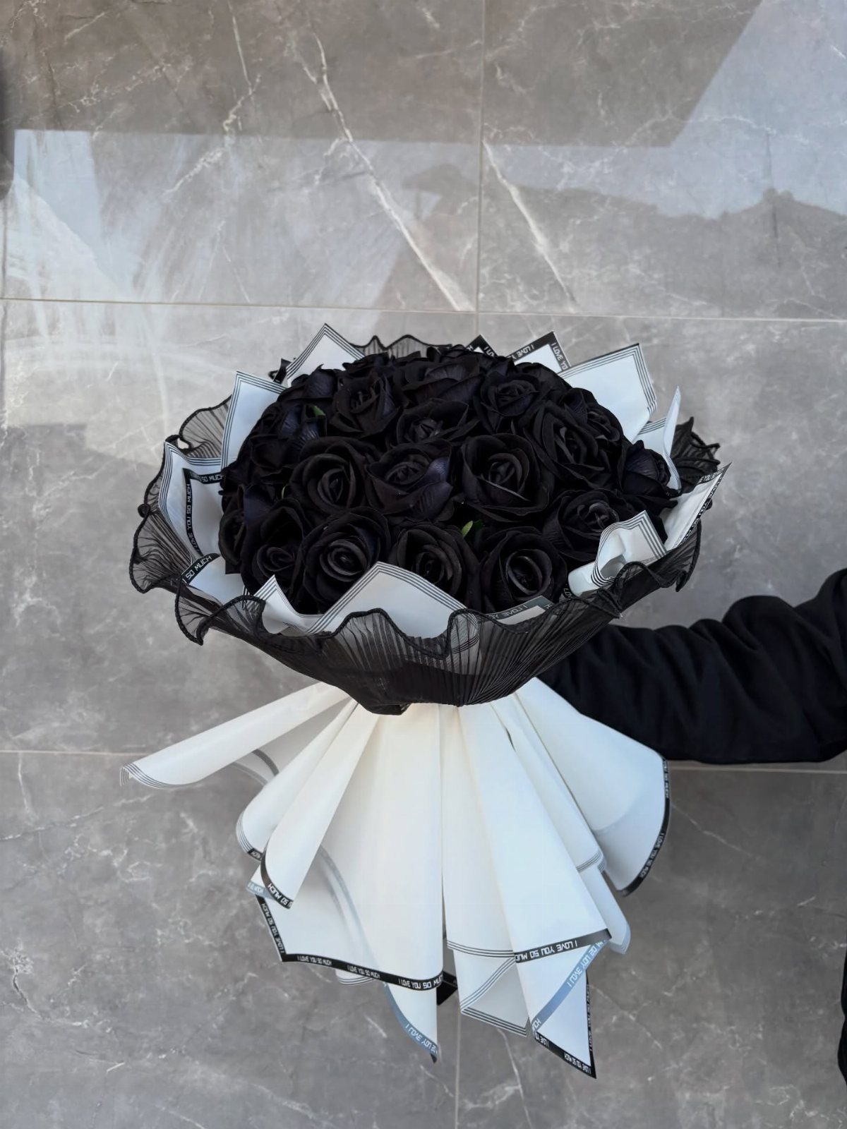 A bouquet of black roses wrapped in white and black decorative paper held against marble.