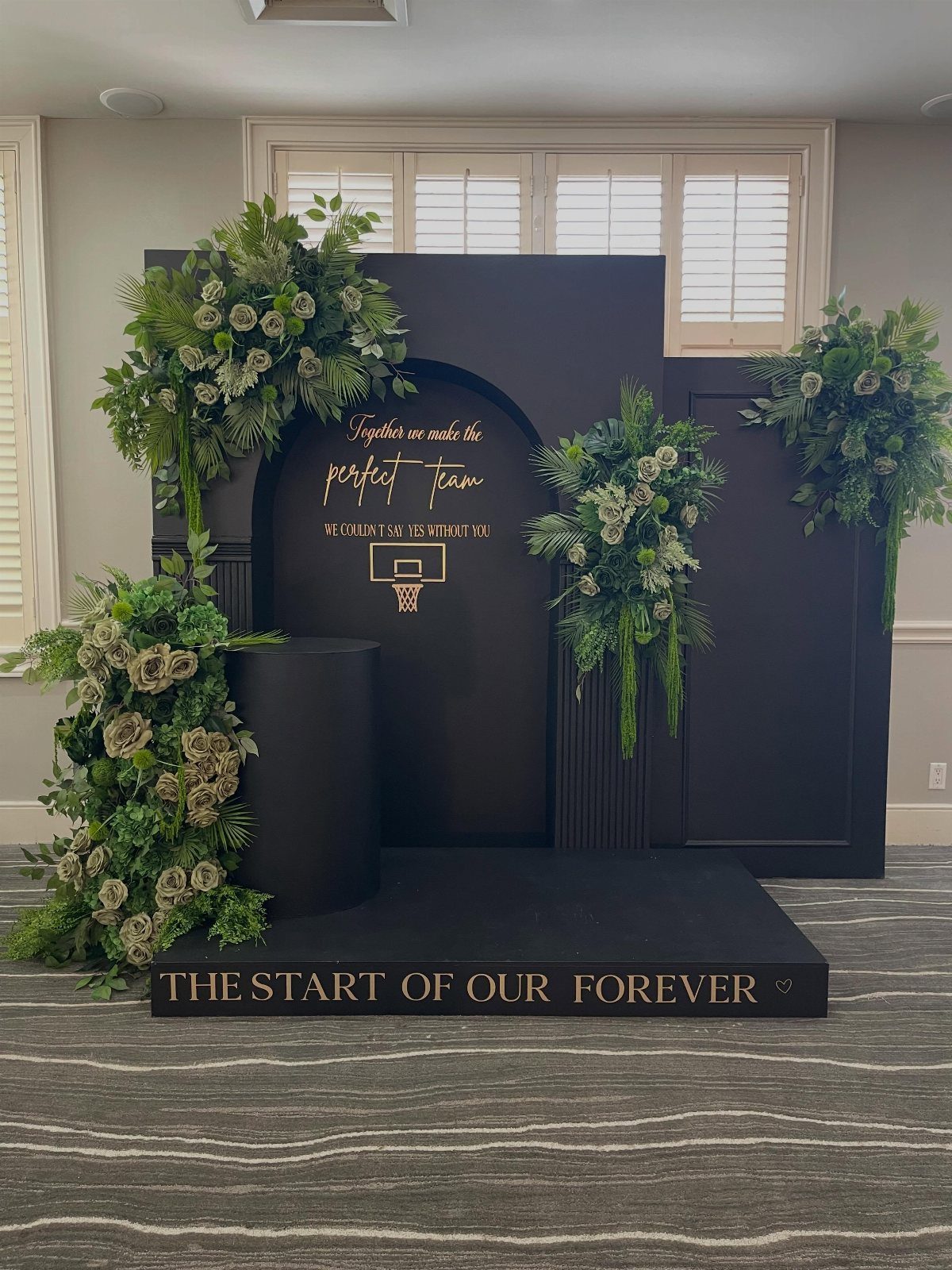 Black wedding backdrop with green floral arrangements and gold calligraphy text on a black platform.