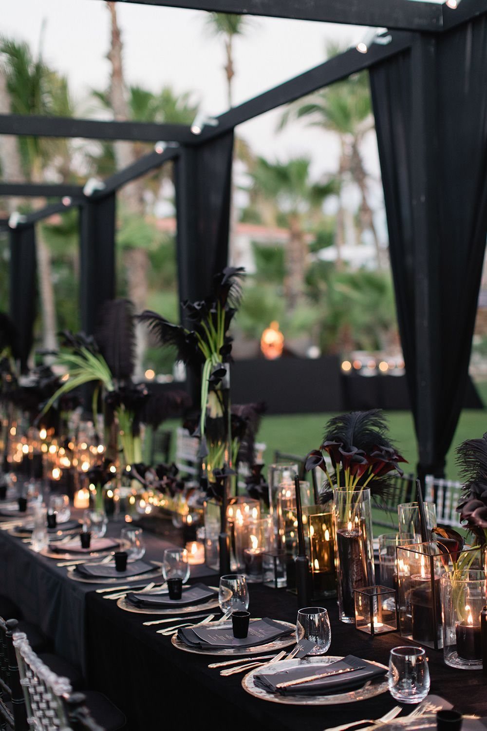 A long wedding reception table featuring black linens, dark floral arrangements, and glowing candles.