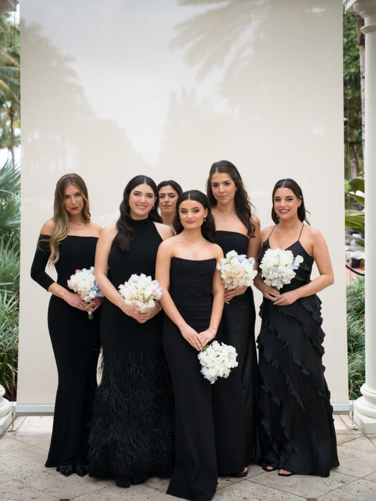 Six bridesmaids in different styles of black floor-length dresses holding white flower bouquets.