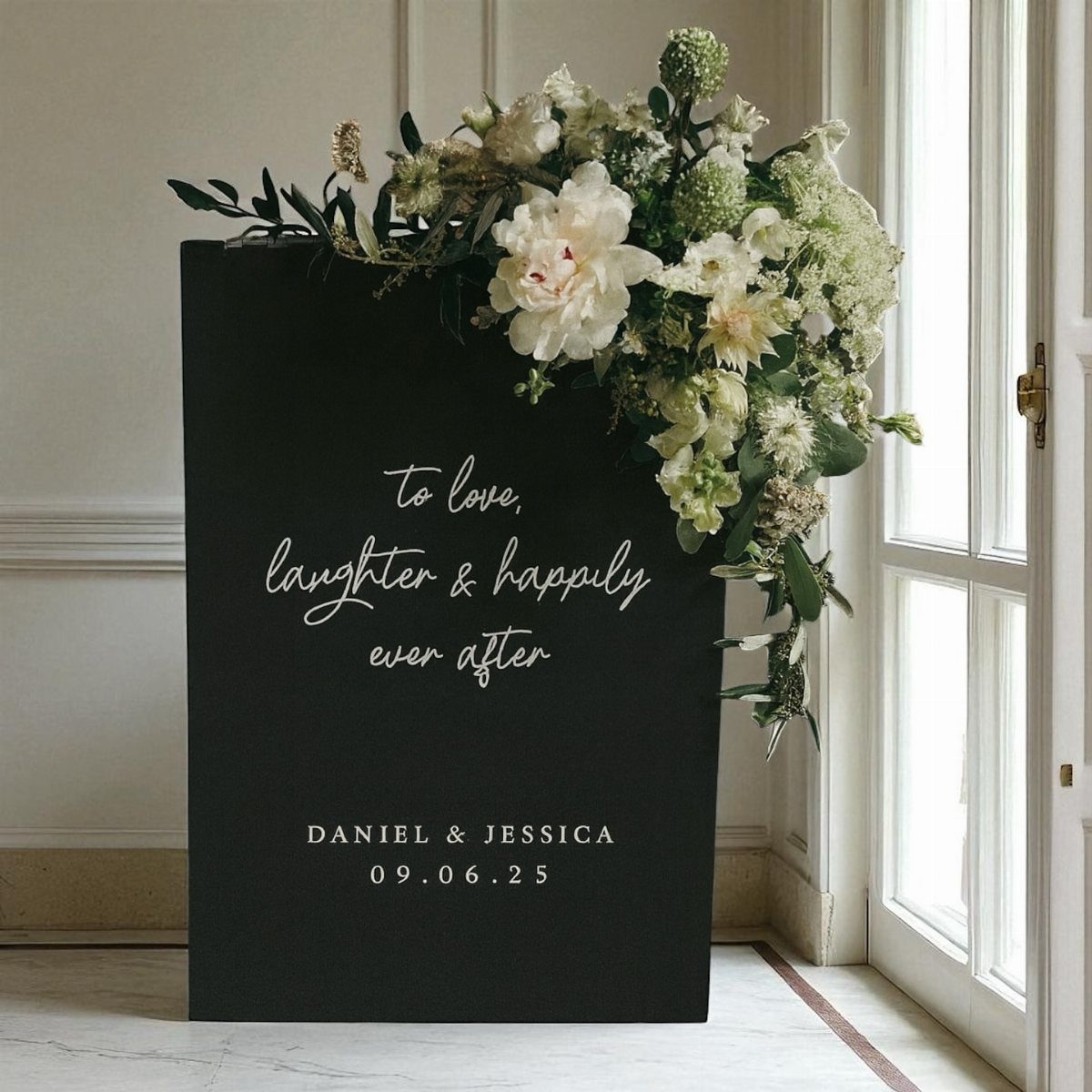 A black wedding welcome sign with white floral arrangements and personalized text standing near a window.