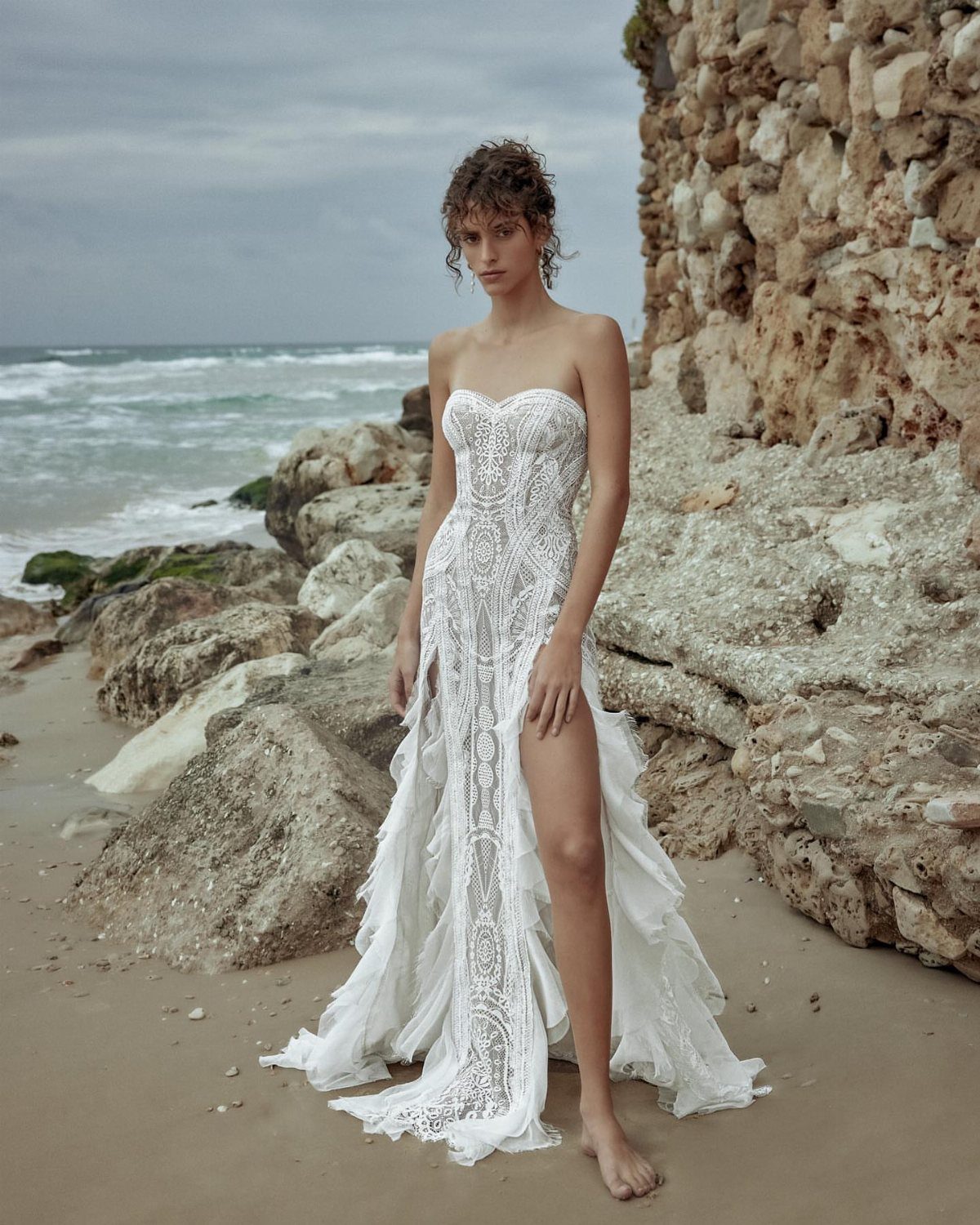 8 Alternative Wedding Dresses for Every Kind of Modern Bride 1 A woman in a strapless white lace wedding dress with a high slit standing on a beach.