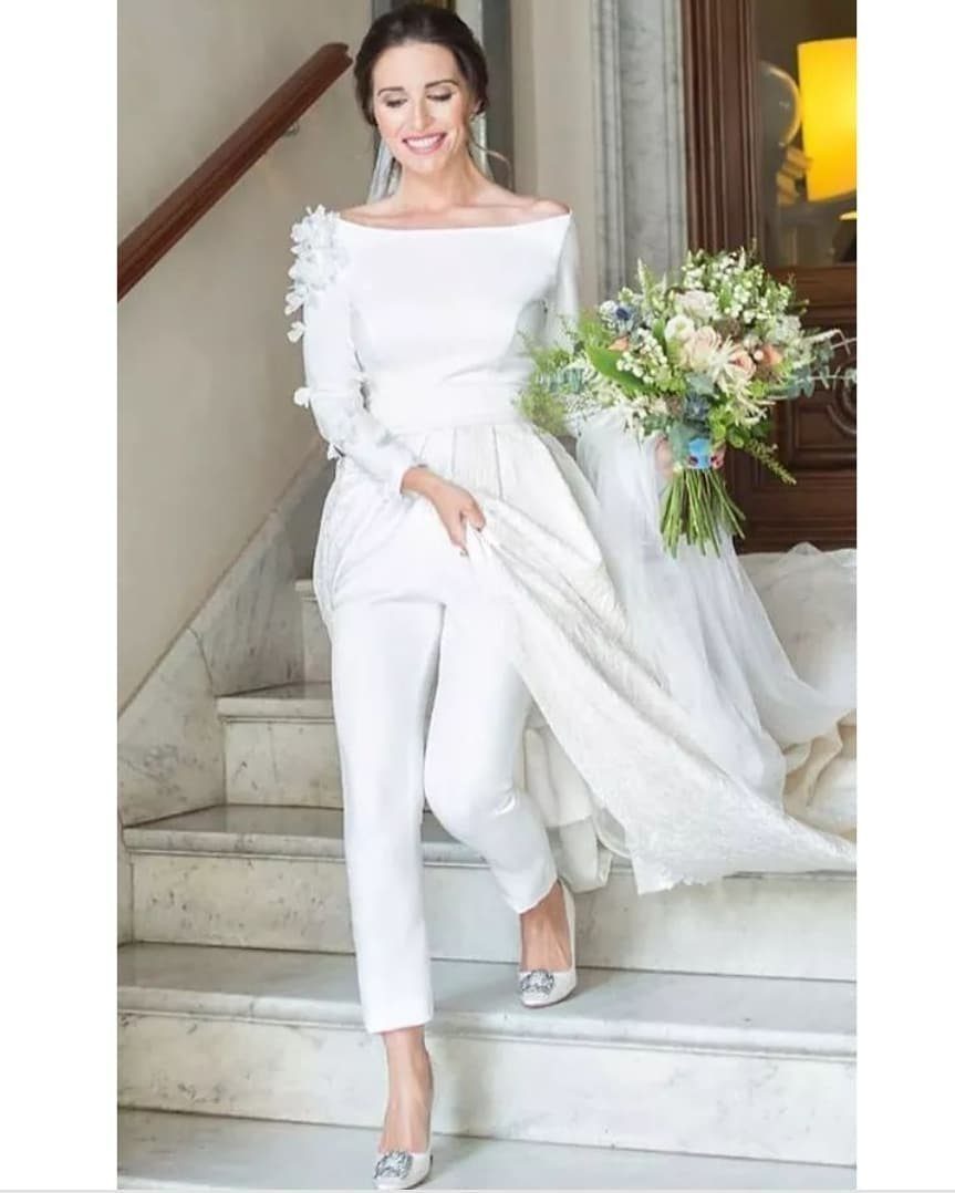 8 Alternative Wedding Dresses for Every Kind of Modern Bride 3 A woman in a white bridal pantsuit with a long train holding a bouquet on stairs.