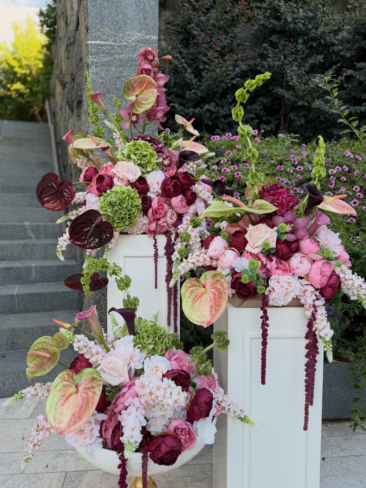 15 Boho Wedding Flowers and Ideas for the Free-Spirited Bride 9 Large pink and burgundy floral arrangements on white pedestals outdoors near a stone staircase.