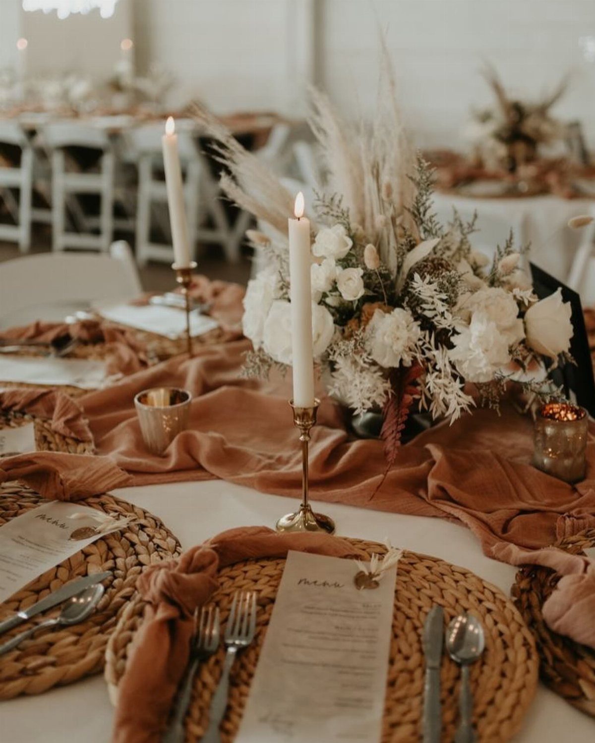 15 Boho Wedding Flowers and Ideas for the Free-Spirited Bride 14 Wedding table decorated with white flowers, pampas grass, taper candles, and terracotta fabric runner.