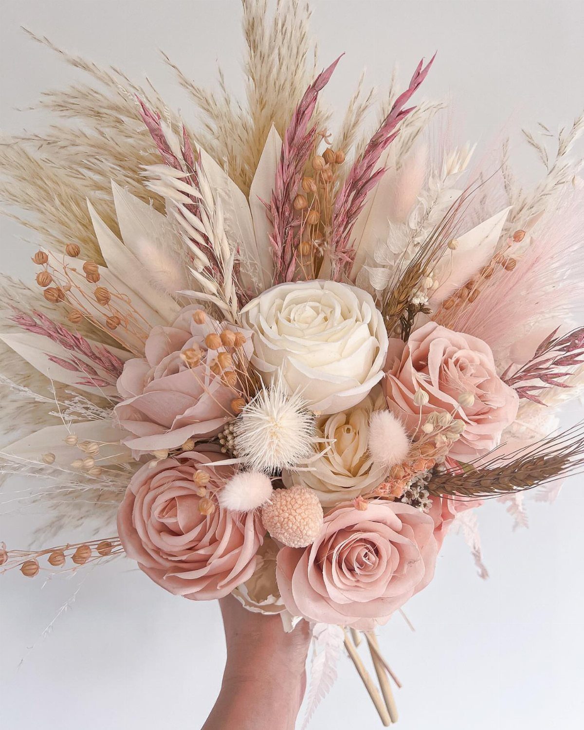 15 Boho Wedding Flowers and Ideas for the Free-Spirited Bride 11 A wedding bouquet with pink roses and dried pampas grass held against a white background.