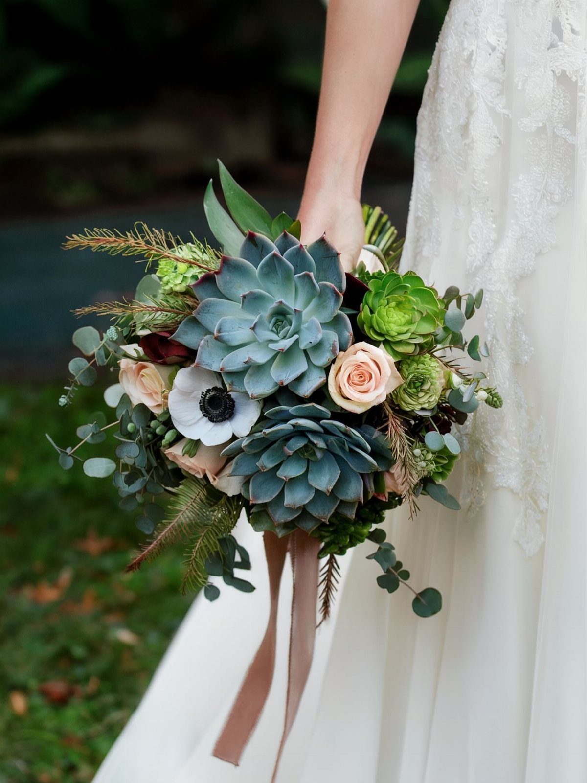 15 Boho Wedding Flowers and Ideas for the Free-Spirited Bride 6 A bridal bouquet containing large succulents, pink roses, and white anemones held against a white lace dress.