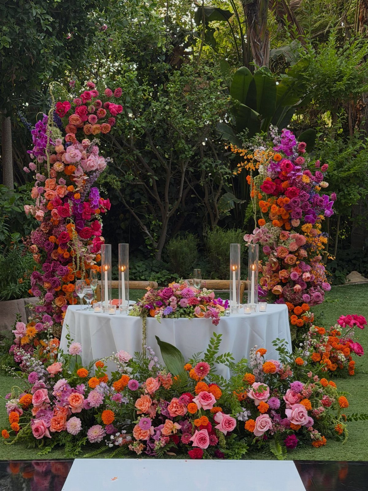 15 Boho Wedding Flowers and Ideas for the Free-Spirited Bride 10 Vibrant pink and orange floral arrangements surrounding a white table with lit taper candles outdoors.