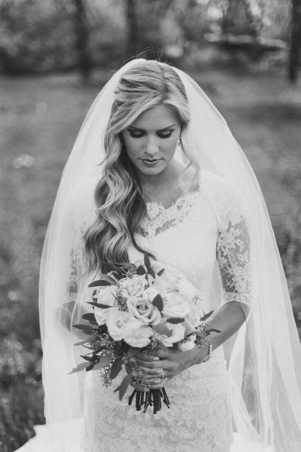 8 Bridal Hair with Veil Ideas for Every Length and Style 3 A bride with side-swept wavy hair, a lace dress, and a long veil holding a rose bouquet.