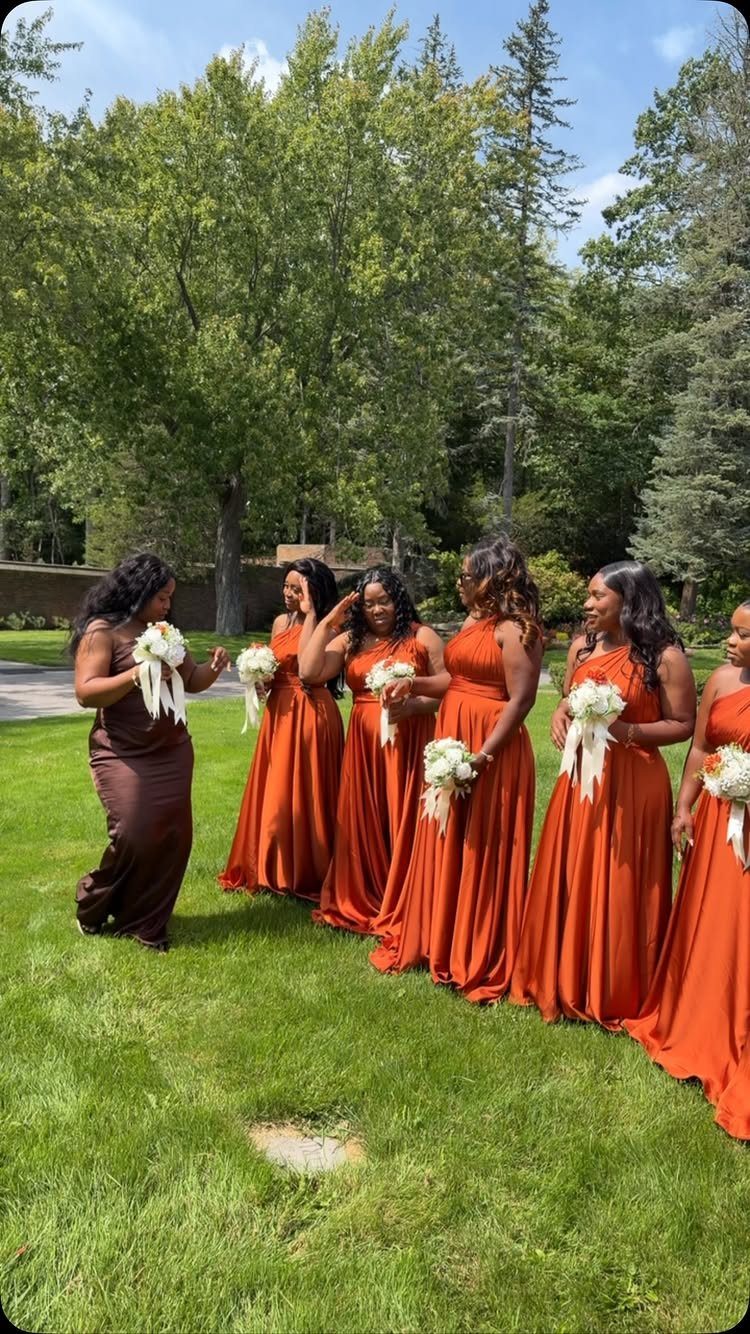 Six bridesmaids in burnt orange long dresses holding white bouquets on a green lawn.