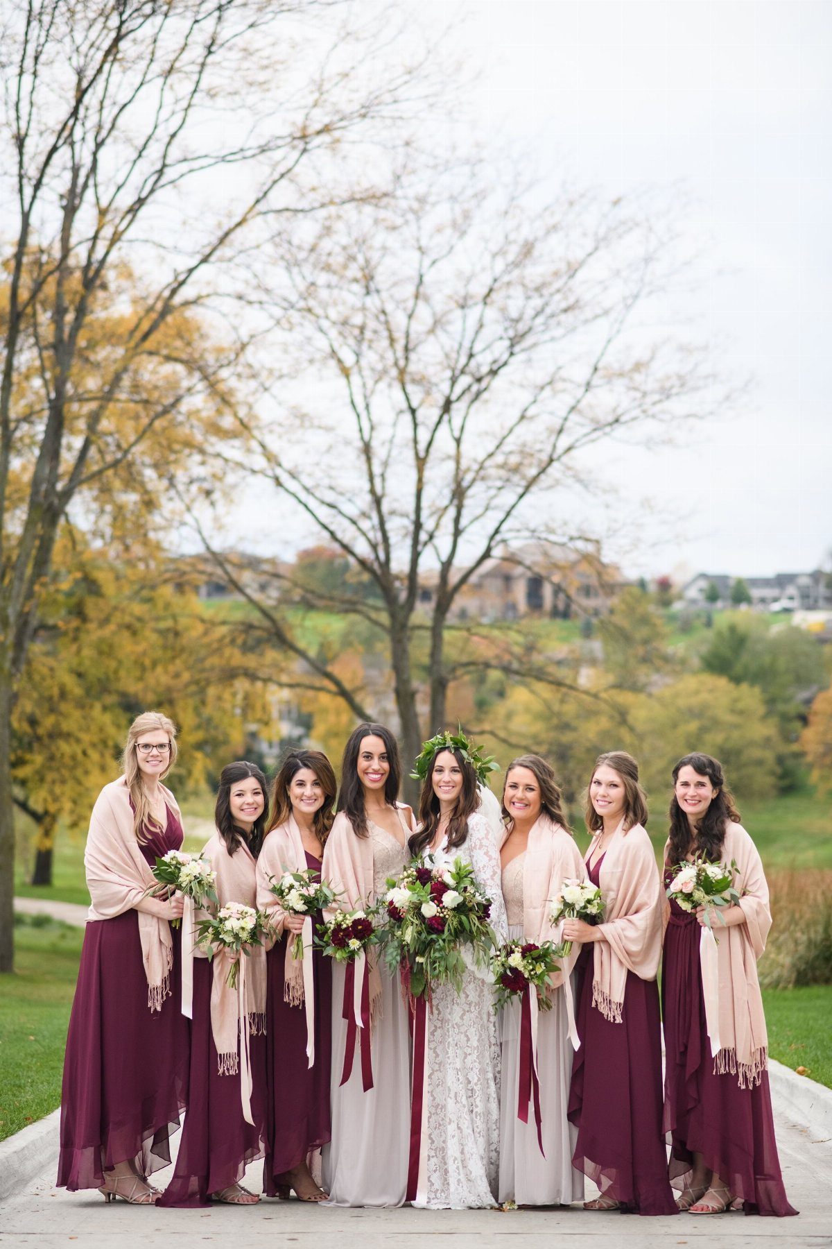 Bridesmaids in long burgundy and cream dresses wearing beige pashmina shawls and holding bouquets outdoors.