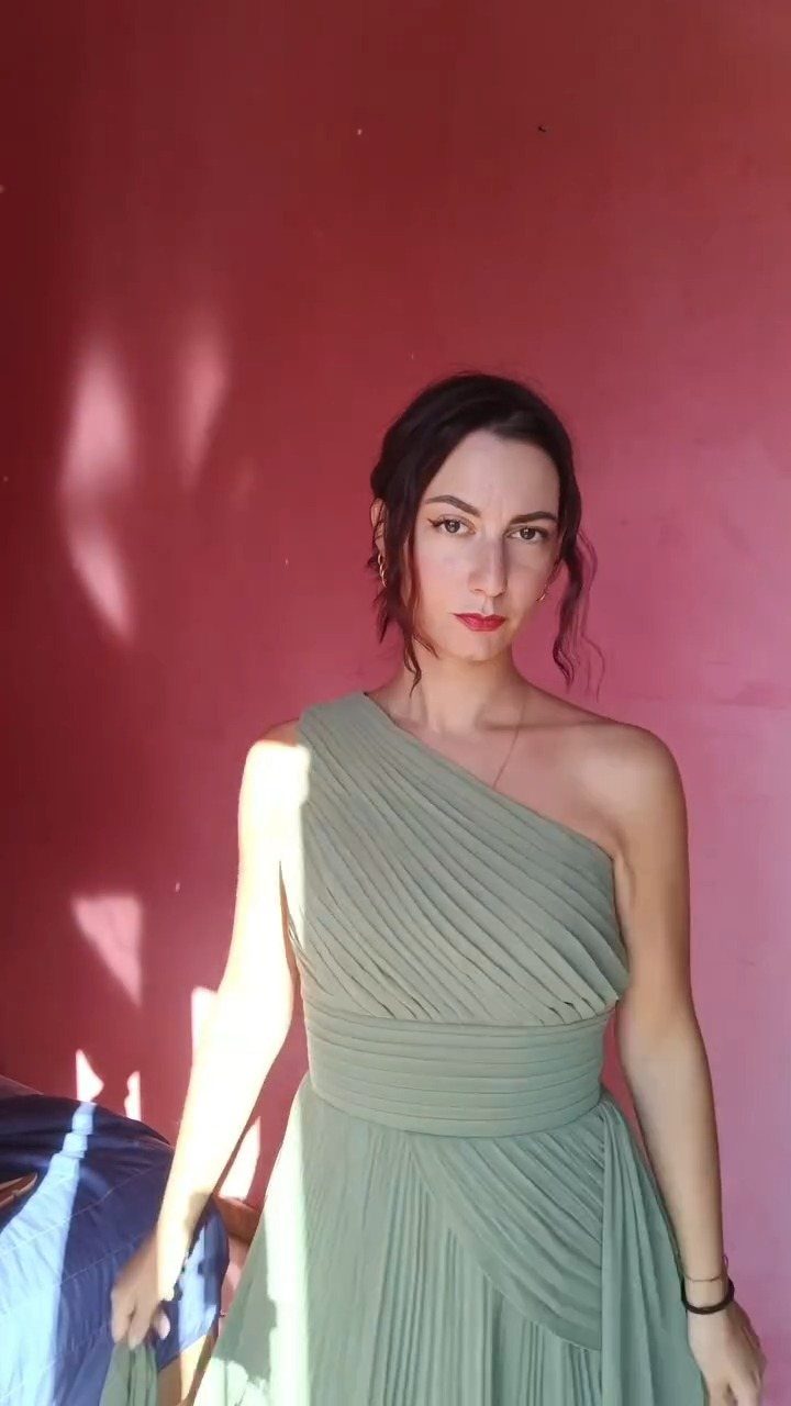 A woman wearing a sage green pleated one-shoulder gown against a pink wall.