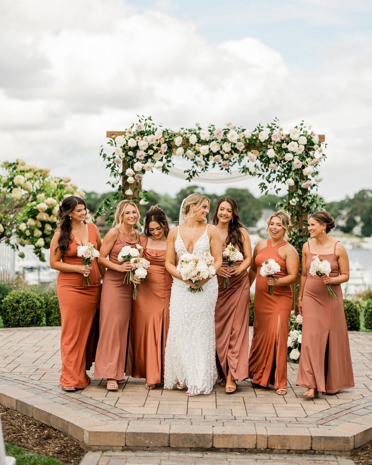 Bridesmaids in terracotta dresses and a bride in white lace stand before a floral arch.