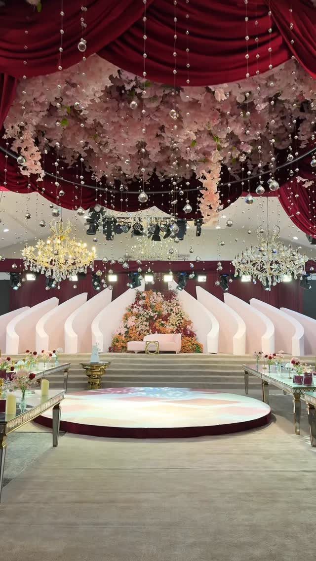 10 Burgundy and Blush Wedding Ideas for Color, Flowers & Decor 9 A grand wedding reception hall with burgundy drapes, pink floral ceiling decor, and a decorated stage.
