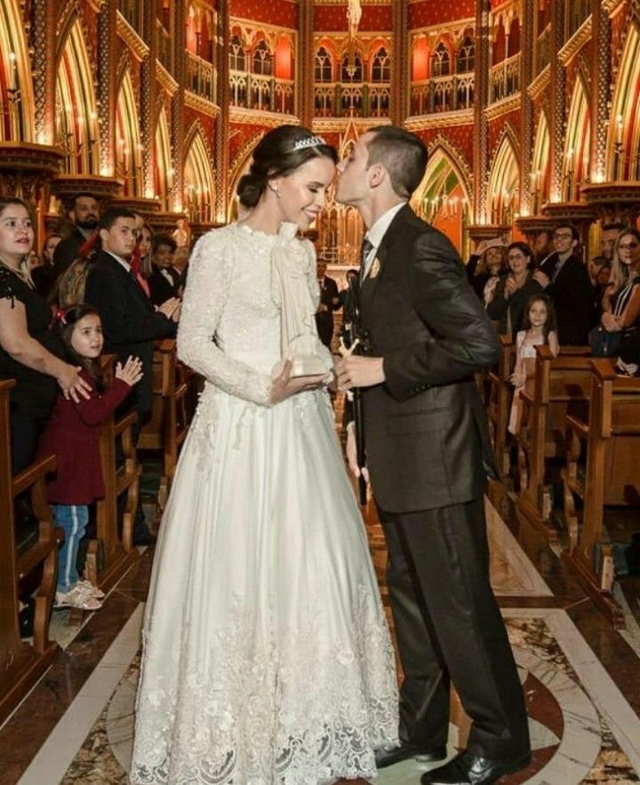 Catholic Wedding Dresses for Modest Church Ceremonies: 6 Ideas 1 A bride wearing a long-sleeved lace gown kisses her groom in a church aisle.