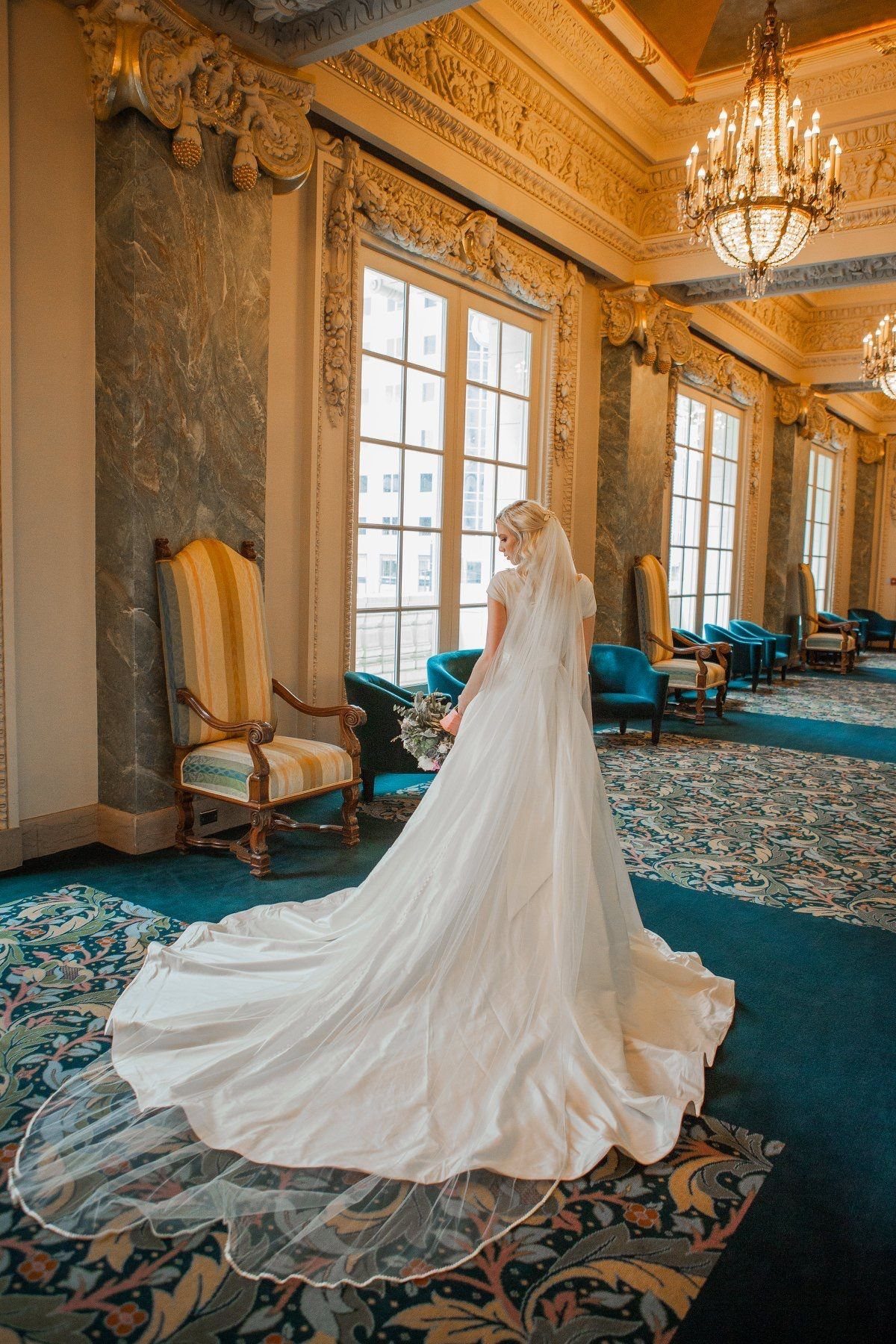Bride in a white wedding dress with a long chapel train standing in an ornate ballroom.