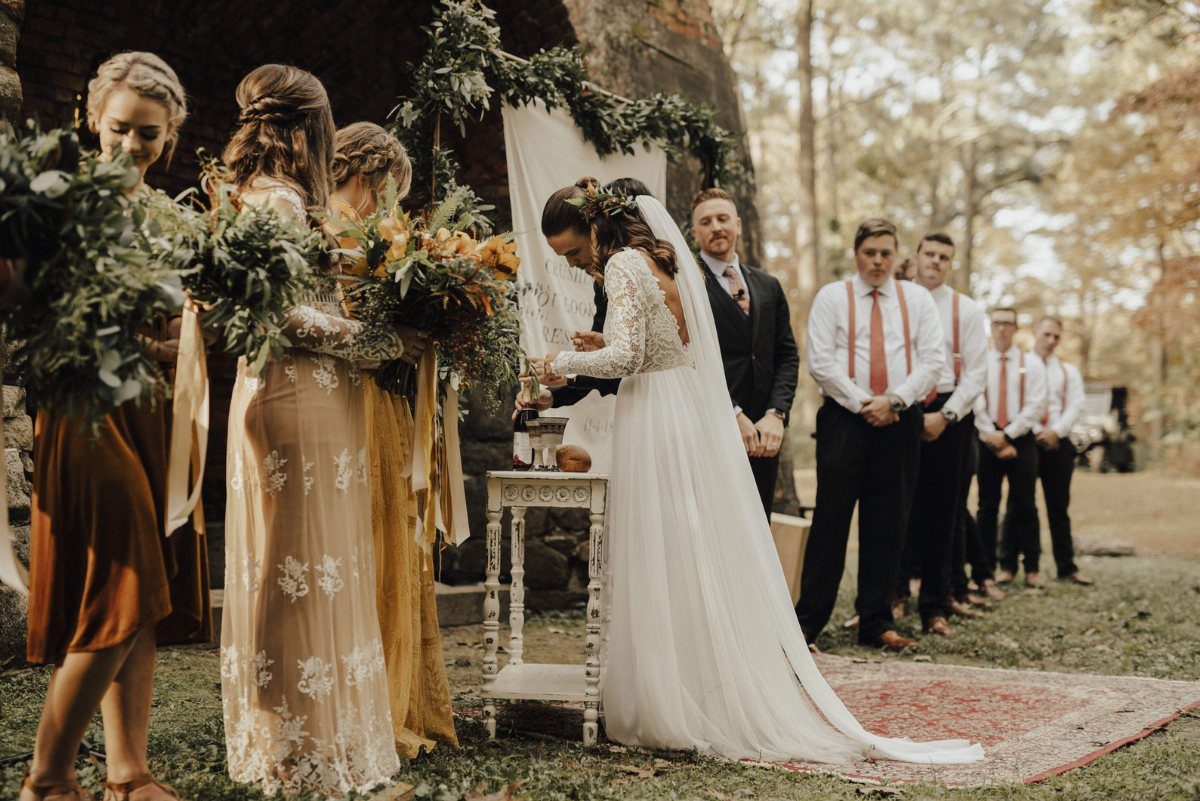 A bride in a white lace wedding dress stands at an outdoor ceremony altar.