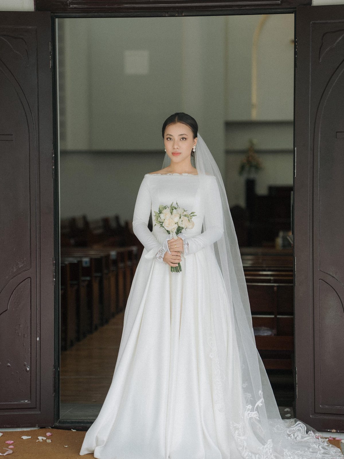 Church Wedding Dress: 10 Styles for the Ceremony 7 A bride in a long-sleeved white satin wedding gown holding a bouquet in a church doorway.