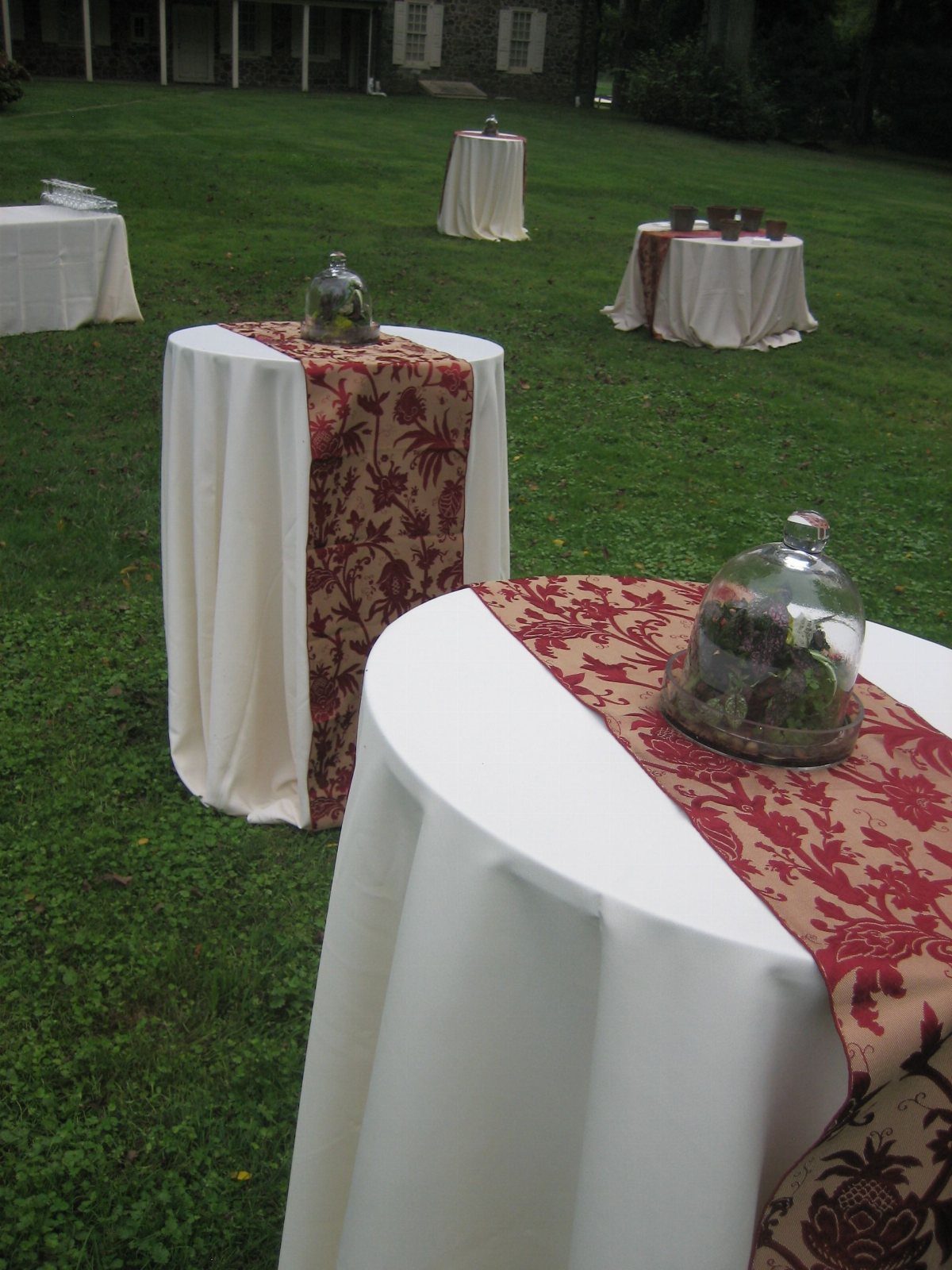 Tall cocktail tables with white cloths and red patterned runners on a green lawn.