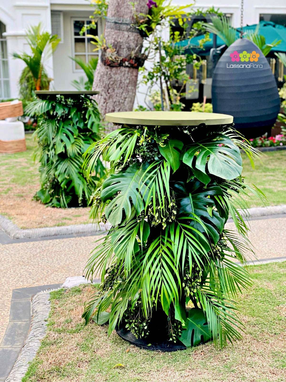 Tropical greenery and monstera leaves cascading down two high cocktail tables outdoors on a lawn.