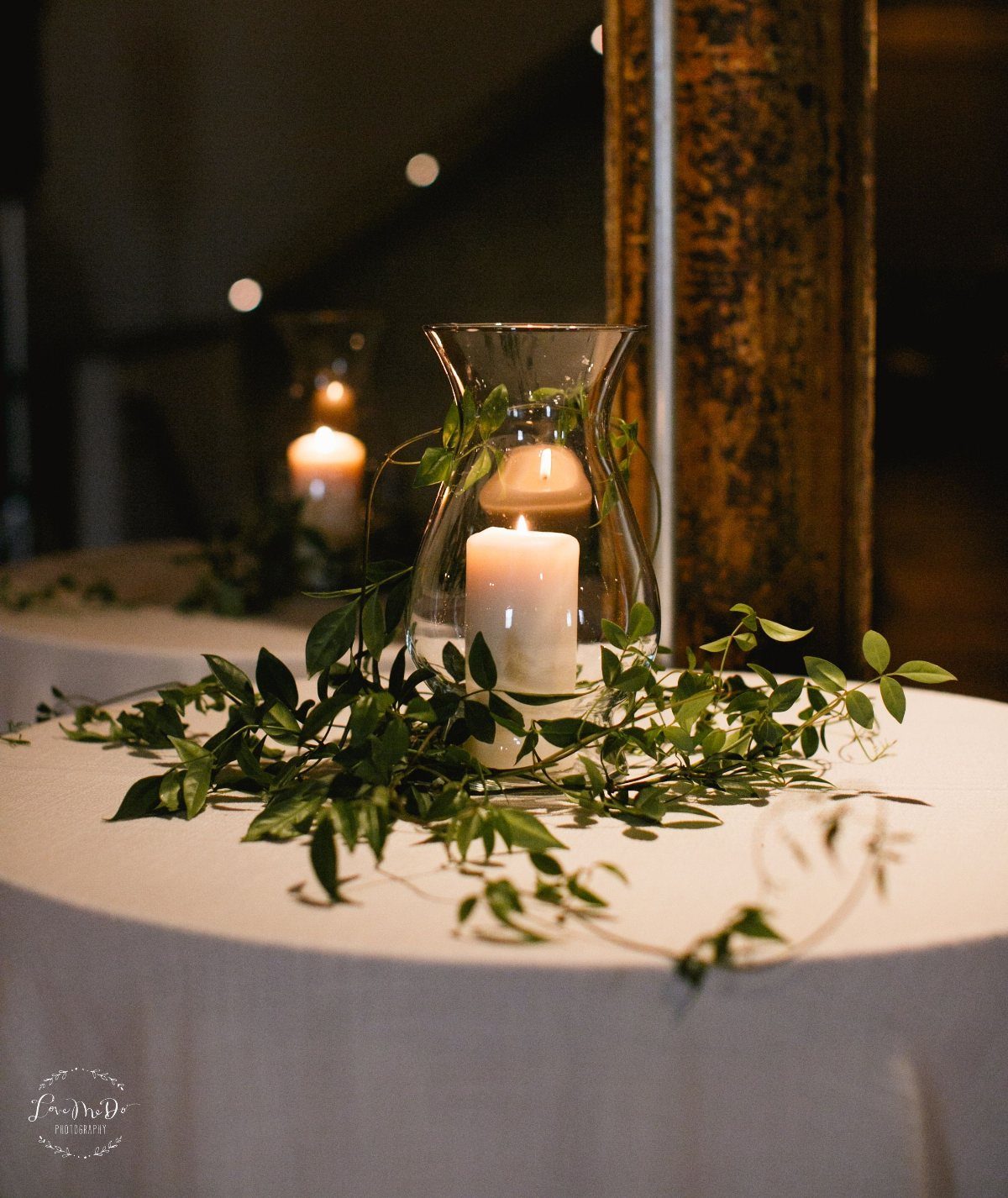 A white pillar candle inside a glass hurricane vase surrounded by green leafy vines on a white table.