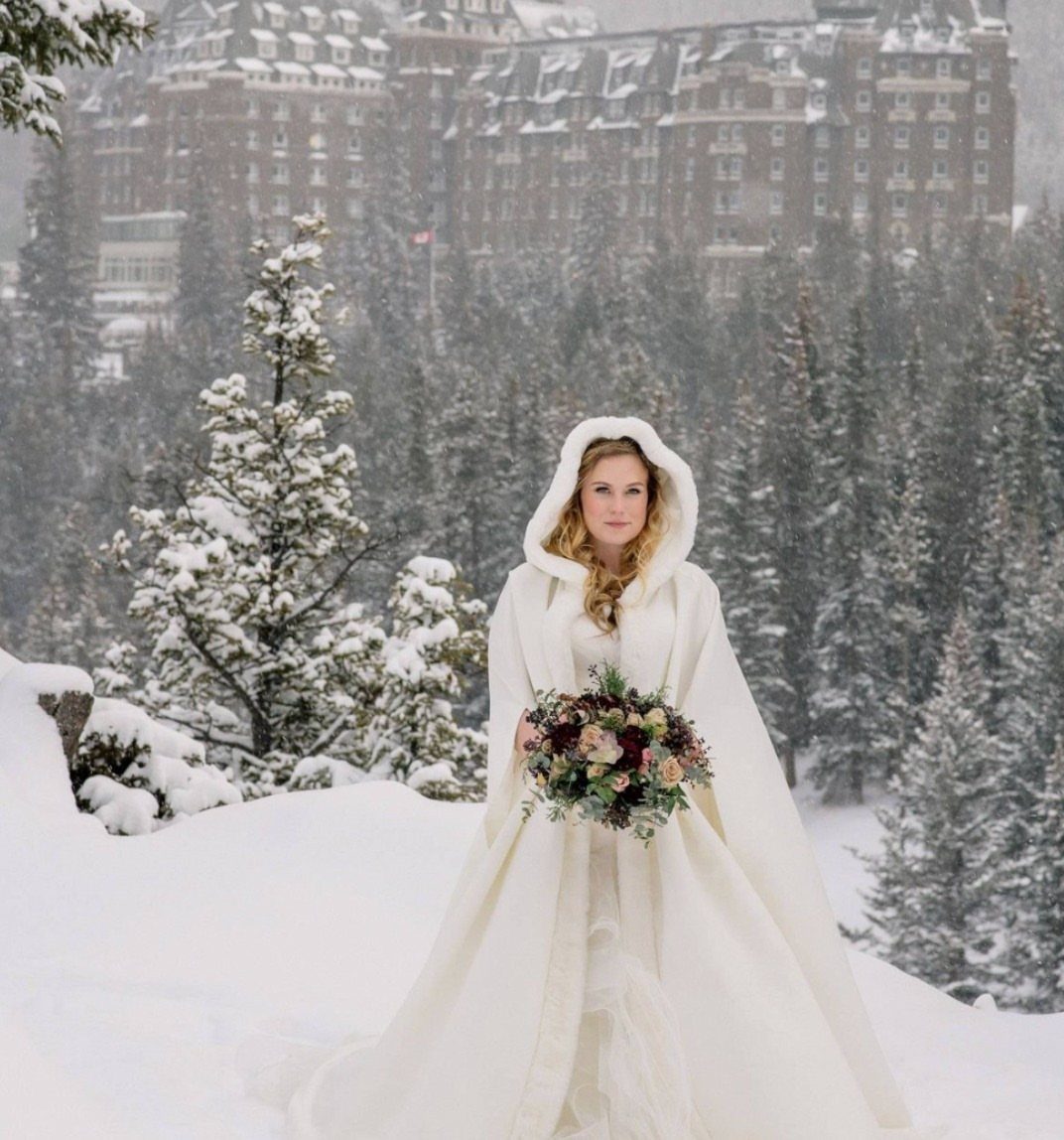 A bride wearing a white wedding gown and a hooded faux fur cape in a snowy forest.