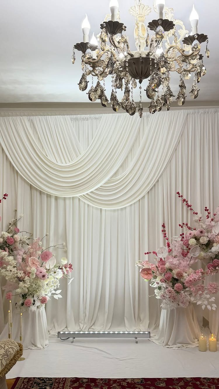 White draped fabric backdrop with pink floral arrangements and a crystal chandelier hanging above.