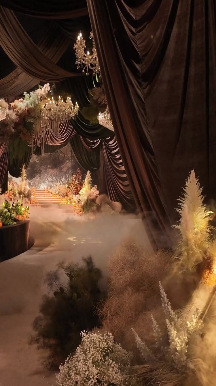 Dark draped fabric and crystal chandeliers illuminate a foggy room filled with white flowers and grasses.