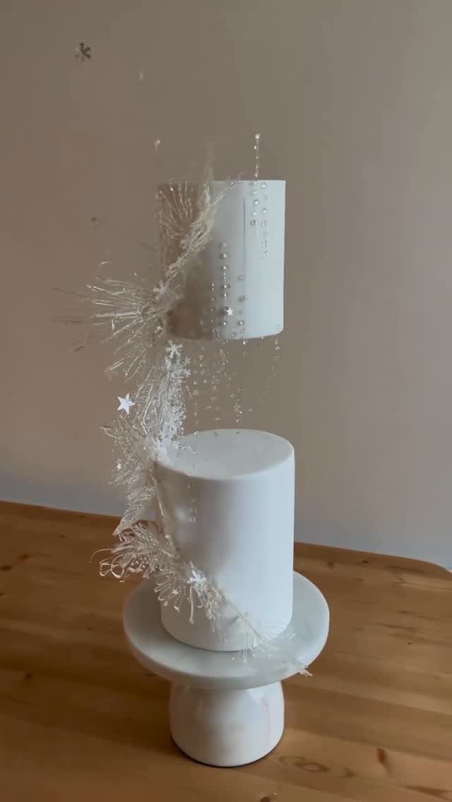 Two-tier white wedding cake decorated with silver glitter and crystalline star ornaments on a marble stand.
