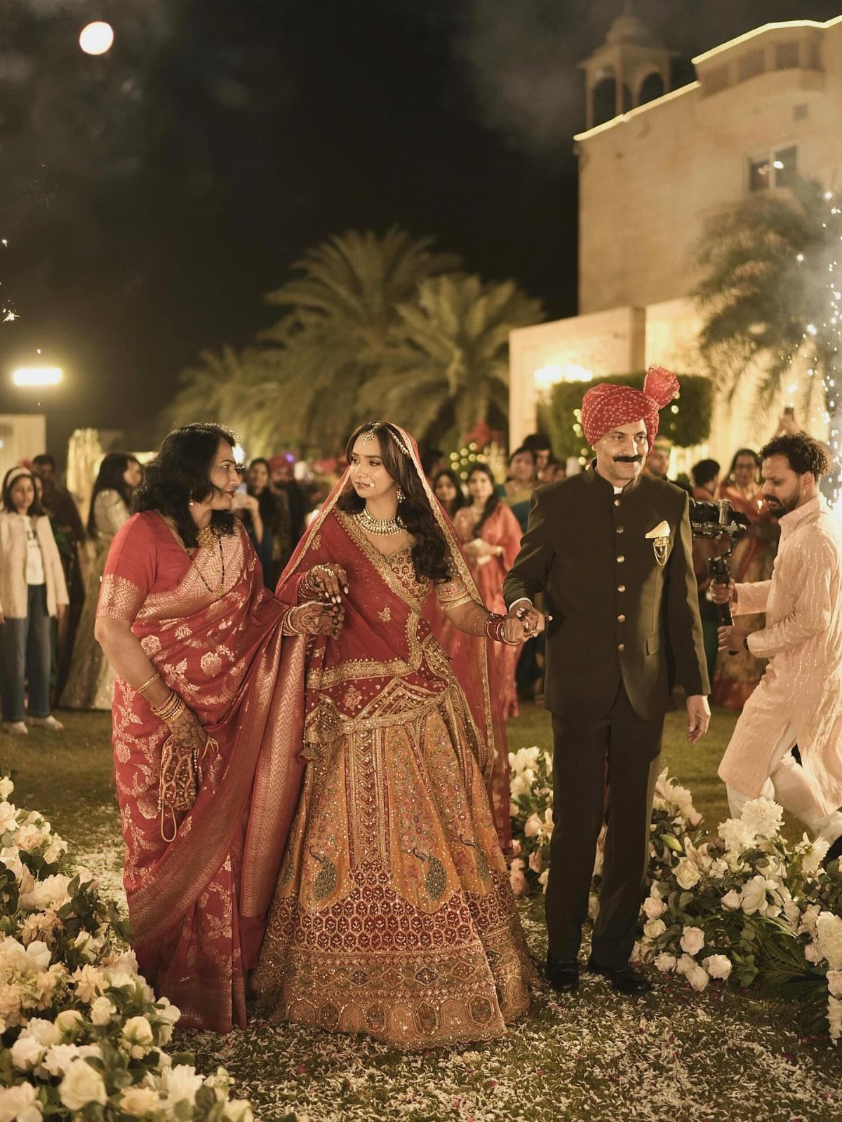 9 Family Wedding Picture Ideas for Every Group 1 A bride in traditional red attire walks between her mother and father outdoors at night.