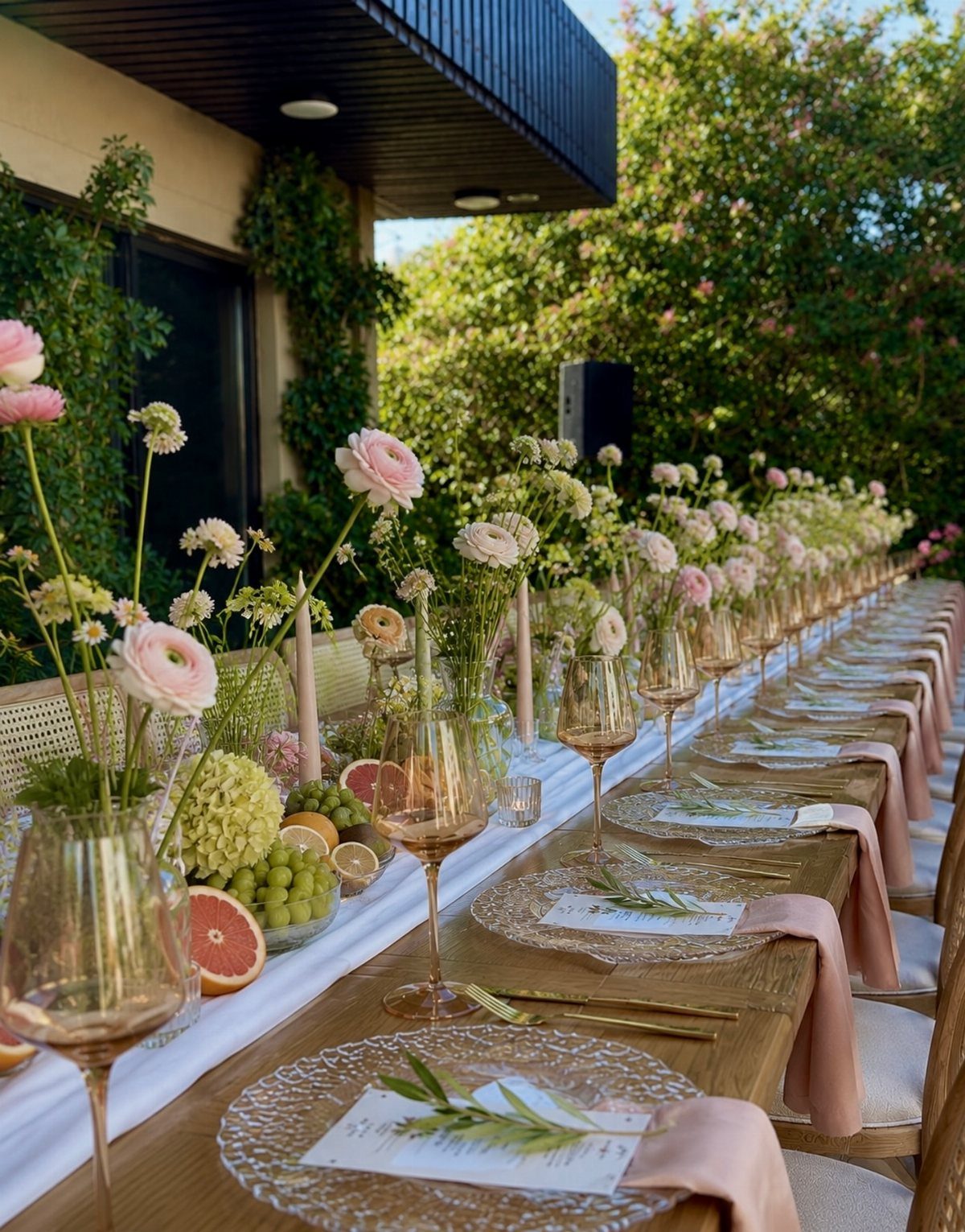A long wooden farm table decorated with pink flowers, glassware, and elegant place settings outdoors.