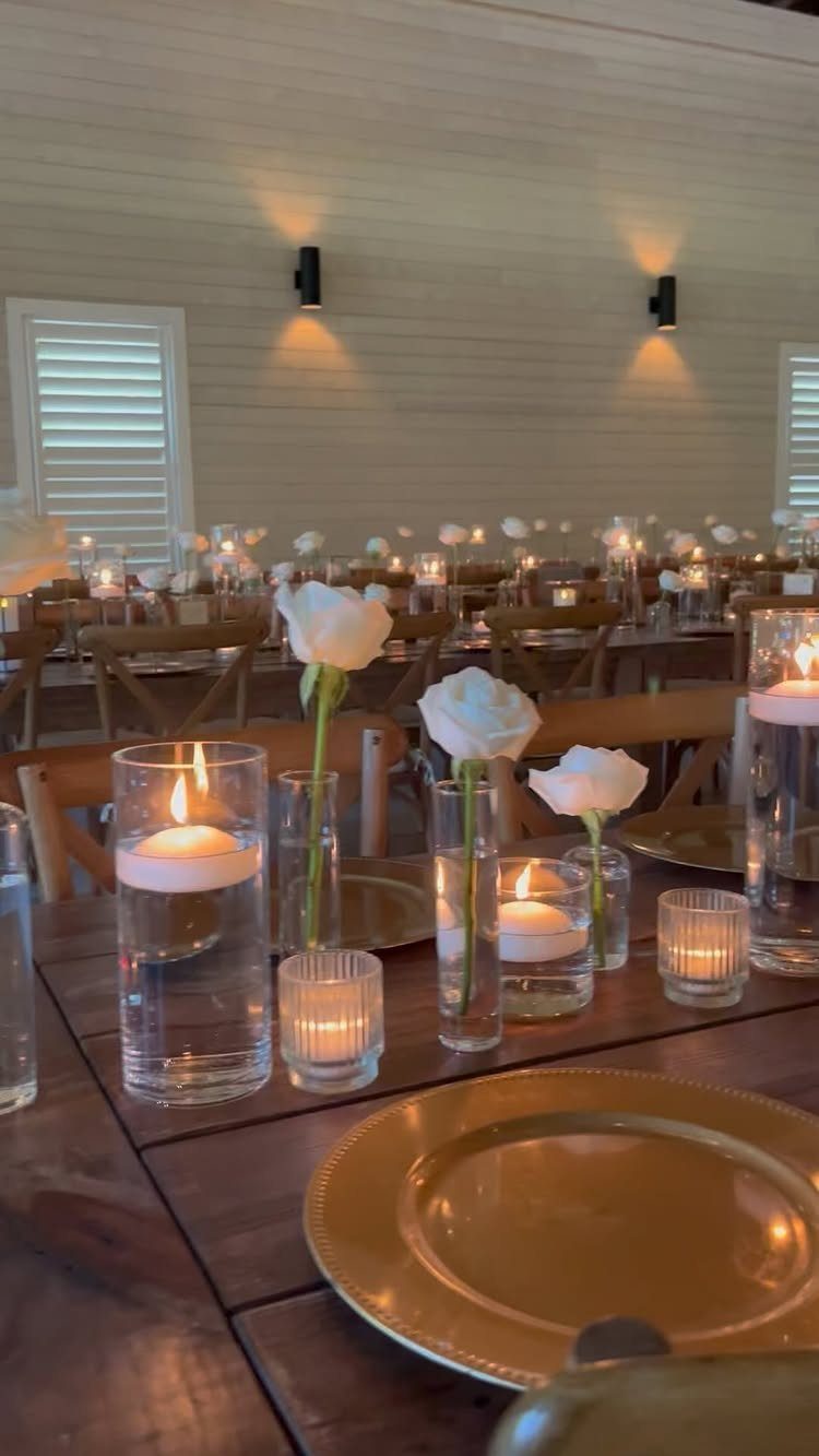 Long wooden farm table decorated with white roses, gold plates, and glowing candles for a wedding.