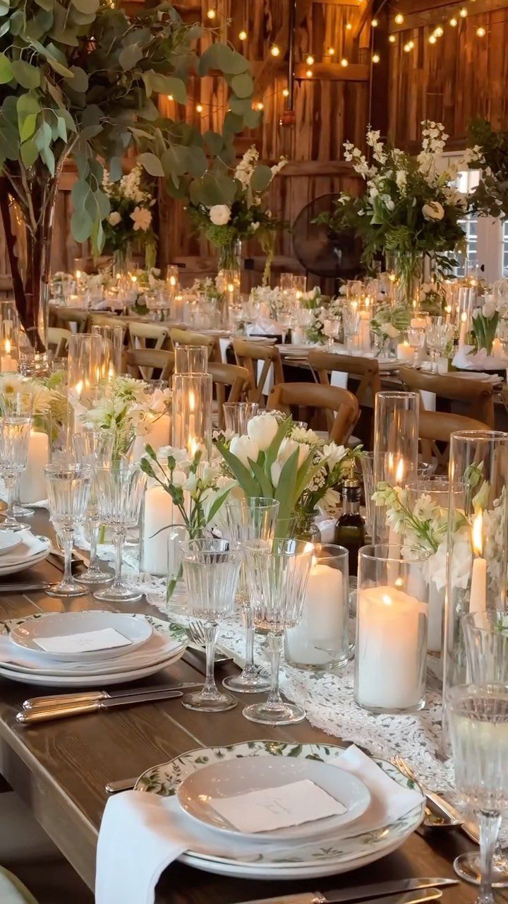 A long wooden farm table decorated with white flowers, greenery, and glowing pillar candles in a rustic barn.