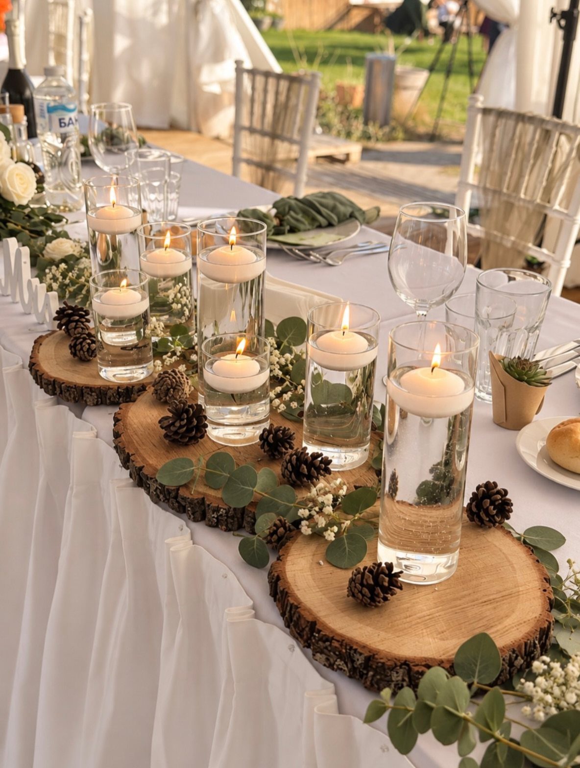 Floating white candles in glass cylinders arranged on wood slices with greenery and pinecones.