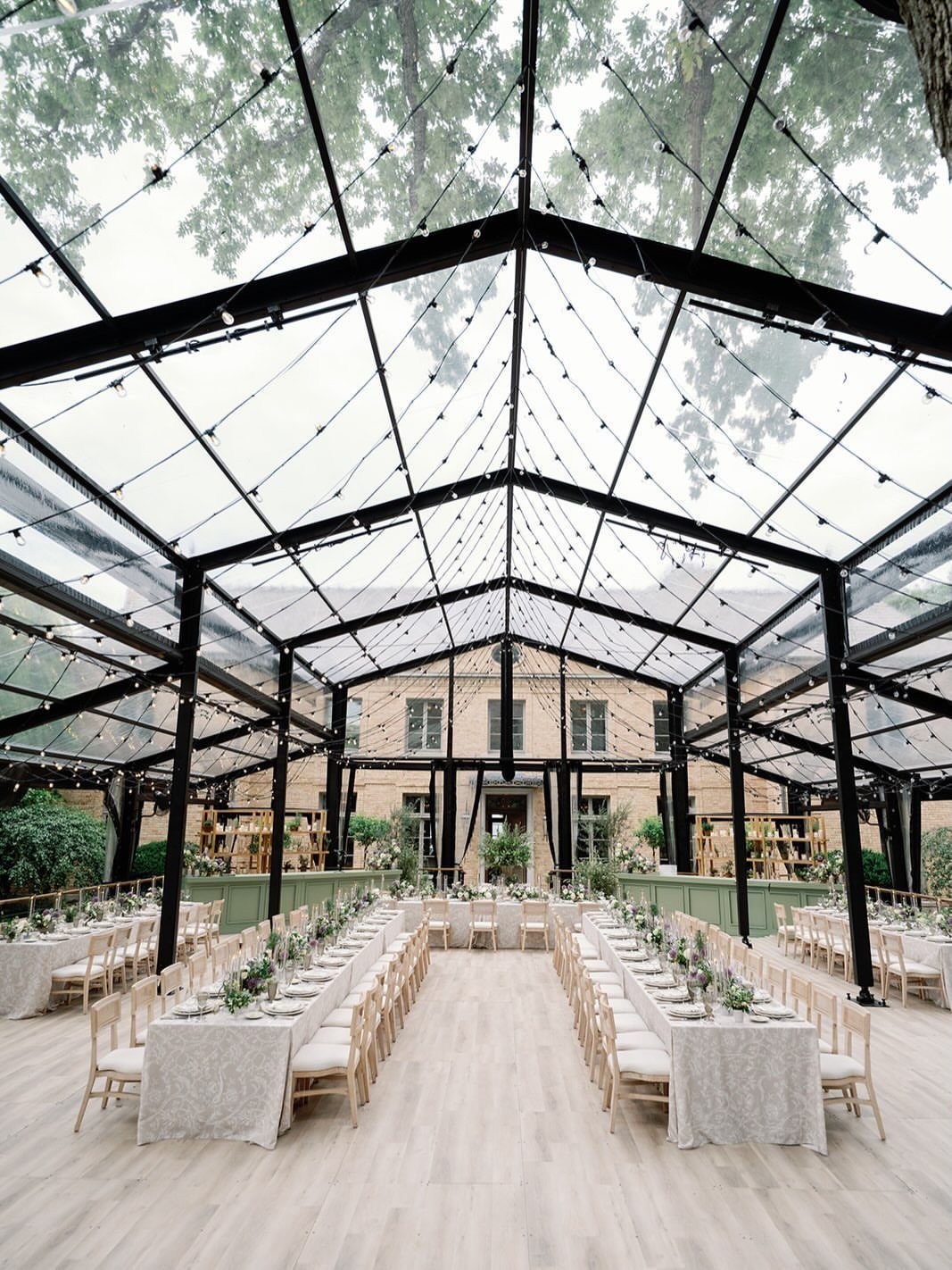 12 Forest Wedding Reception Ideas Under Lanterns and Light 4 Long banquet tables with white linens set under a transparent glass marquee in a forest.