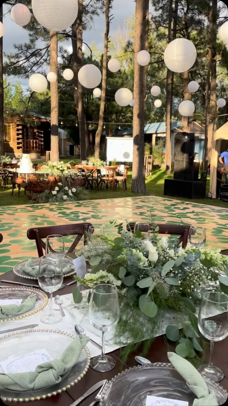 12 Forest Wedding Reception Ideas Under Lanterns and Light 9 A decorated wedding reception table with greenery and glassware set in a forest under hanging lanterns.