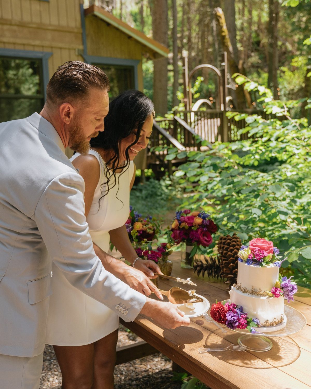 12 Forest Wedding Reception Ideas Under Lanterns and Light 11 A couple cutting a floral wedding cake on a wooden table in a lush forest setting.