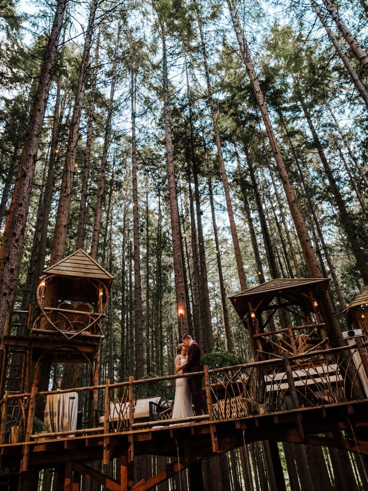 12 Forest Wedding Reception Ideas Under Lanterns and Light 2 A bride and groom embrace on an elevated wooden walkway surrounded by tall pine trees and treehouses.