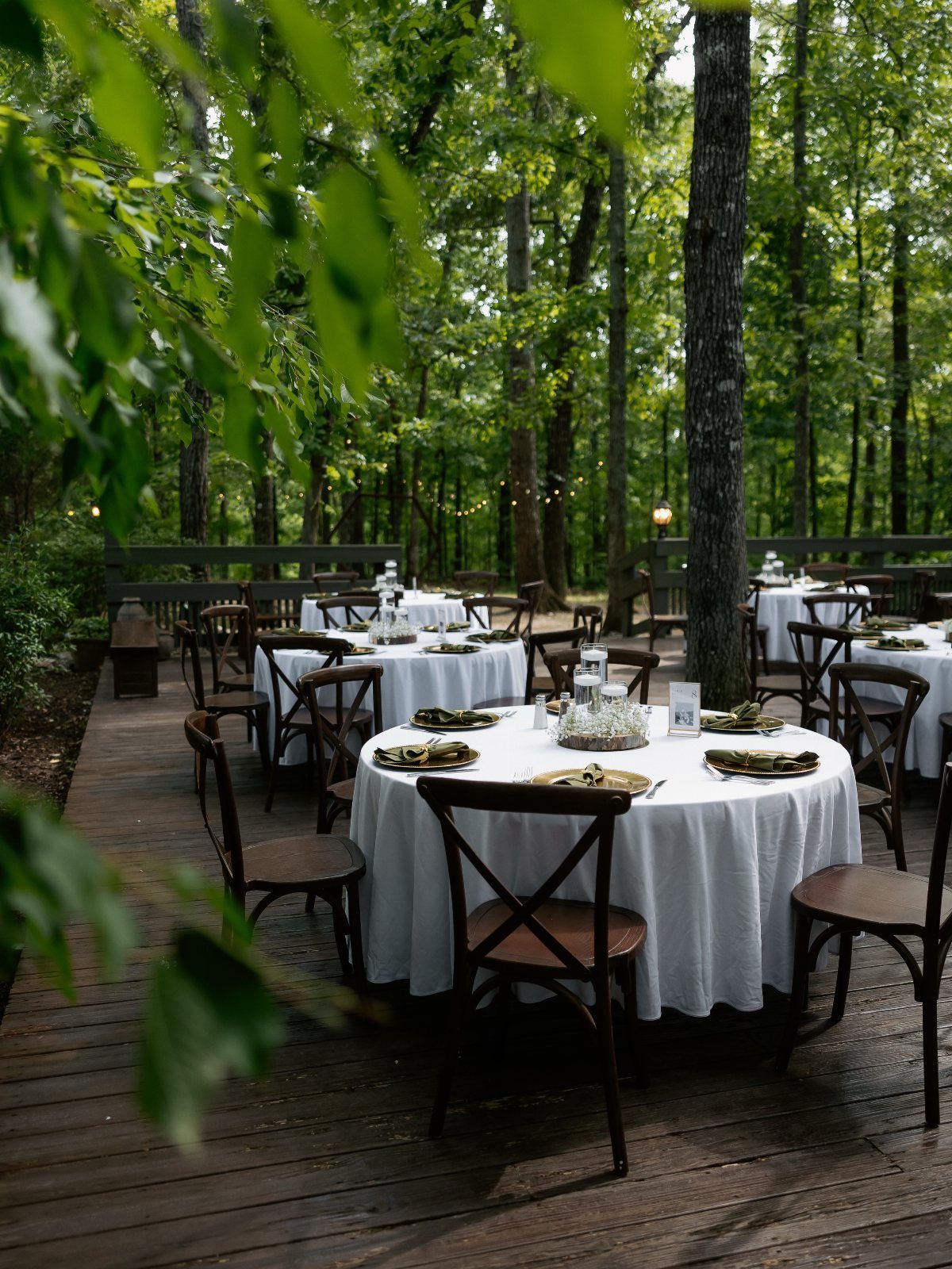 12 Forest Wedding Reception Ideas Under Lanterns and Light 7 Set dining tables with white linens and gold chargers on a wooden deck surrounded by dense green forest trees.