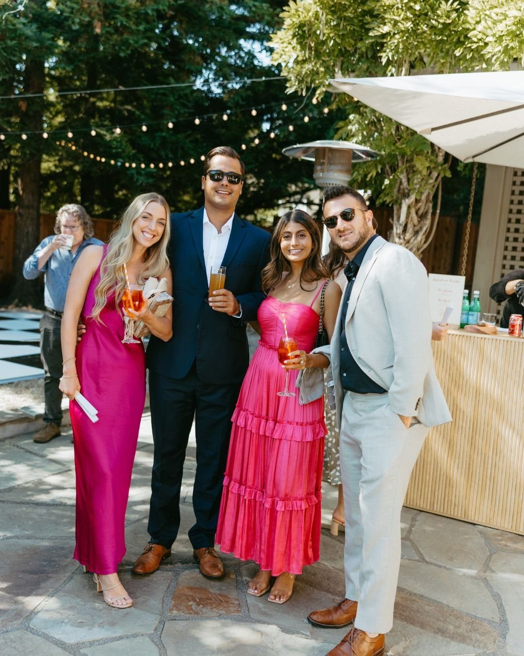 Four people in formal attire posing together at an outdoor garden party with drinks.