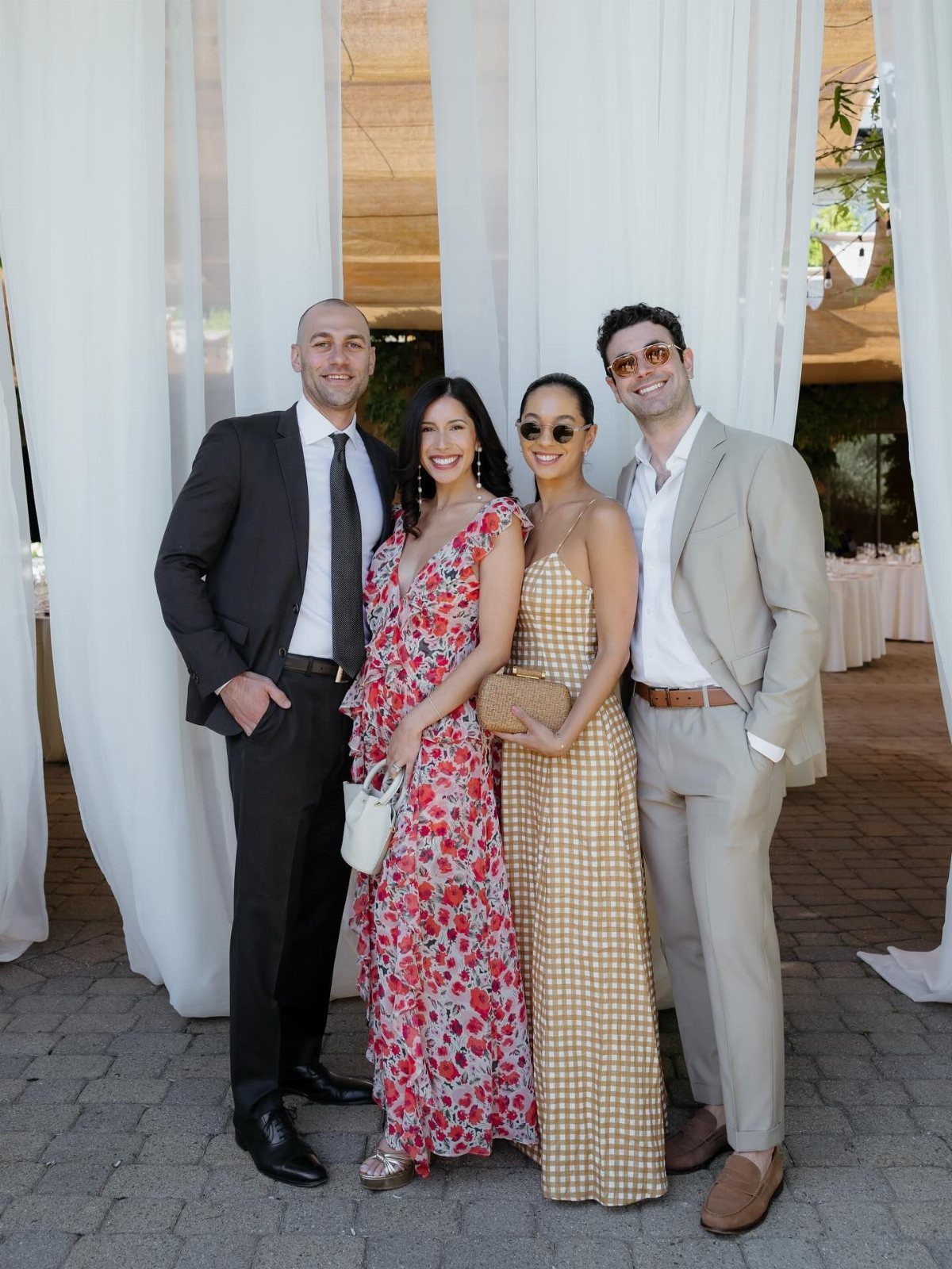 Four people dressed in formal wedding guest attire posing together outdoors against white curtains.