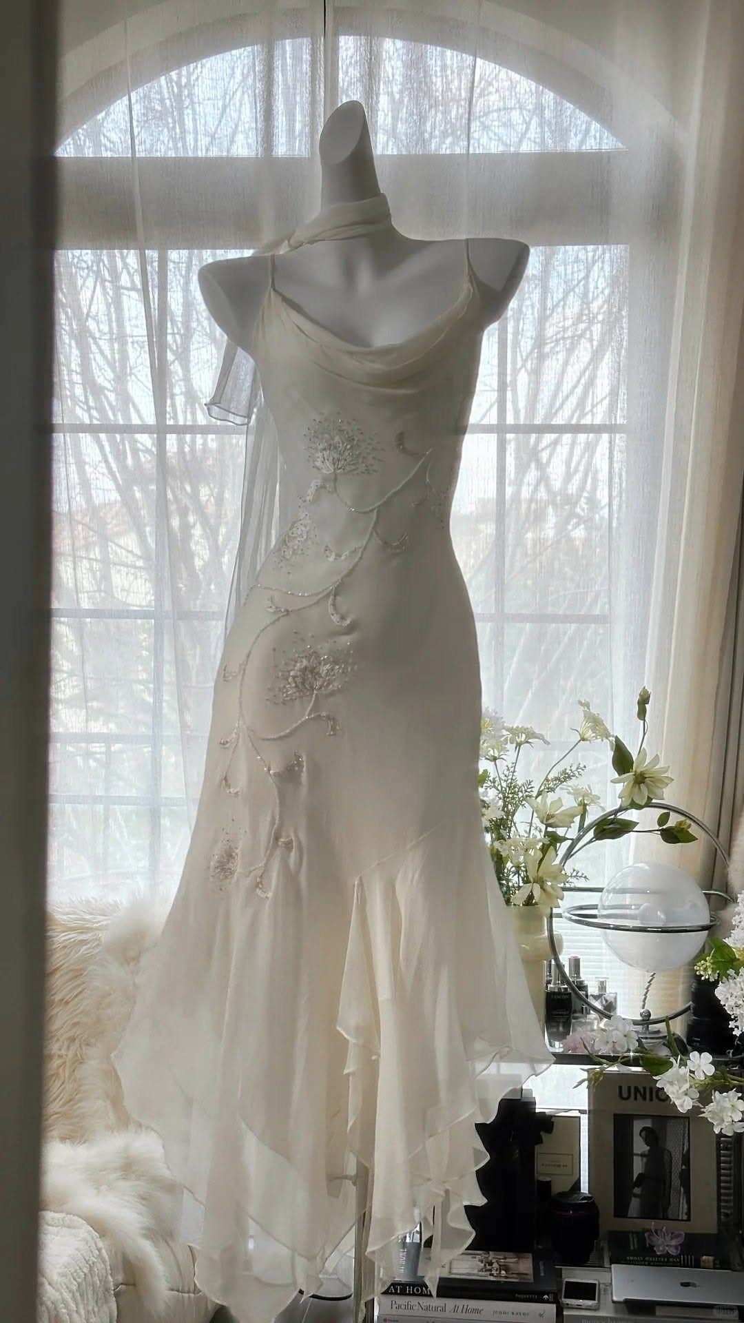 An ivory chiffon wedding dress with floral embroidery displayed on a mannequin by a window.