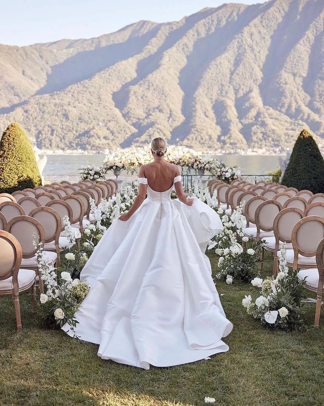 A bride in a white wedding dress with a long train stands in a garden aisle.