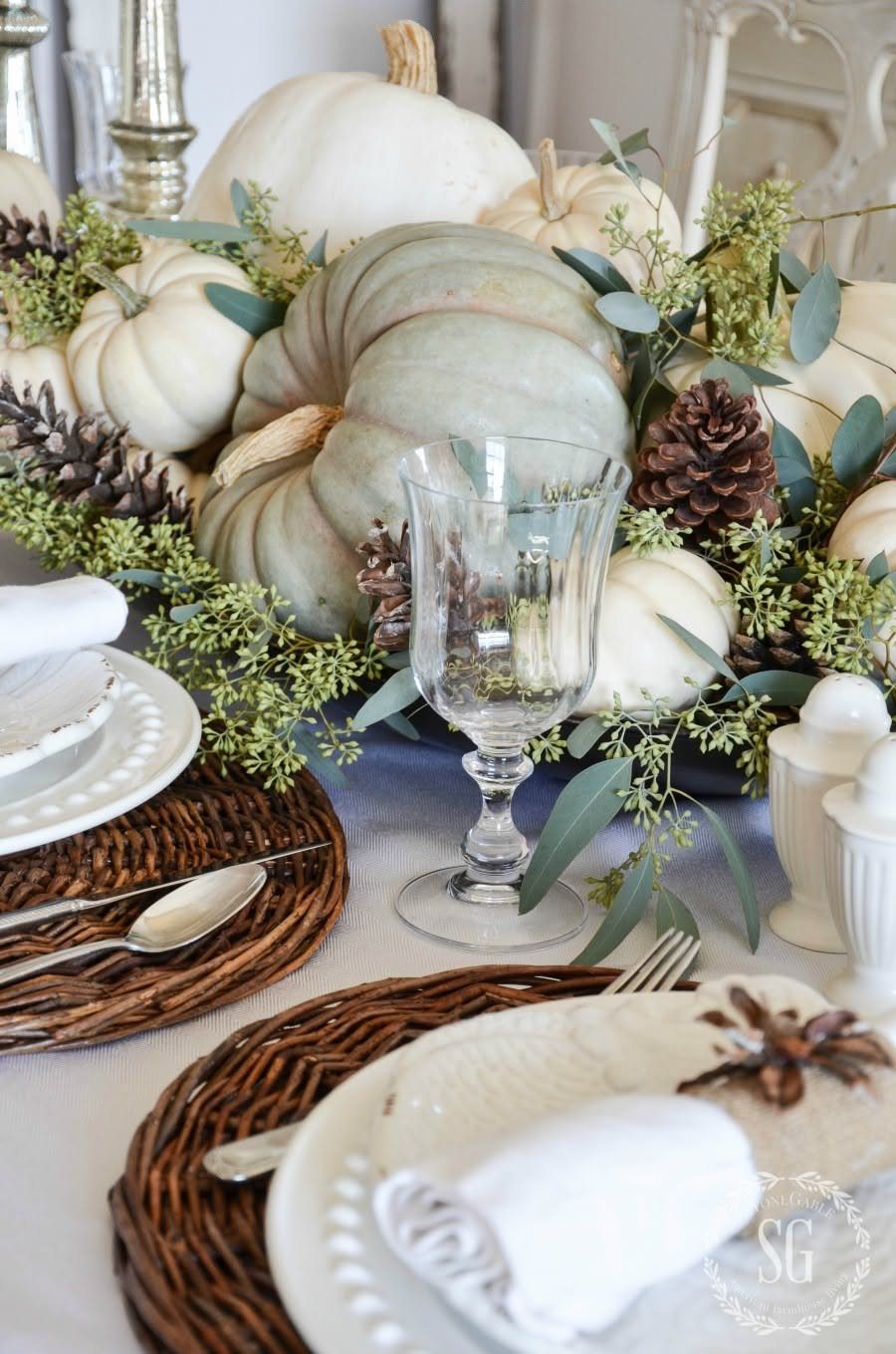 White and green pumpkins mixed with eucalyptus leaves and pinecones arranged as a centerpiece on a table.