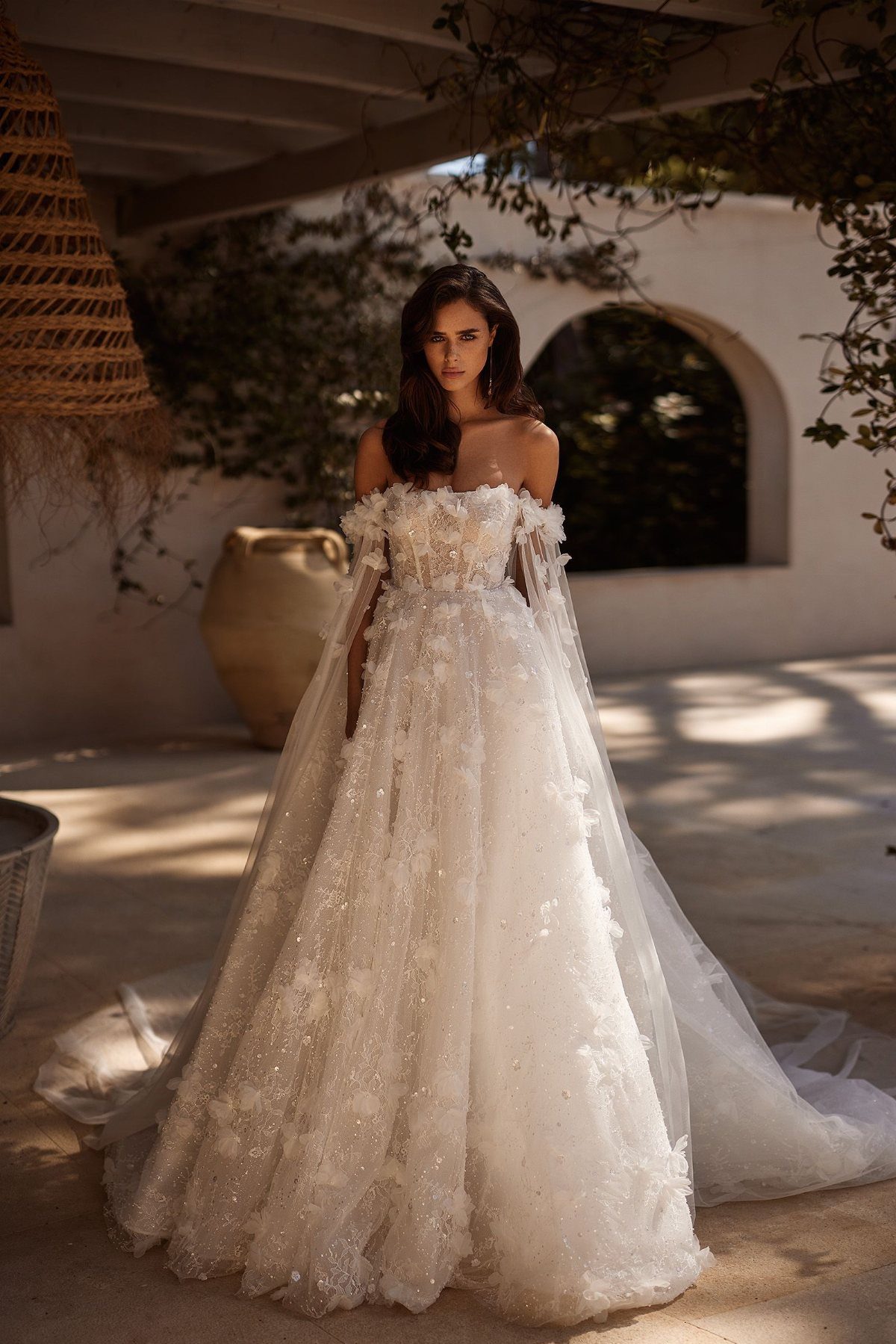 Woman wearing a white off-the-shoulder wedding dress with 3D floral embellishments and sheer cape sleeves.