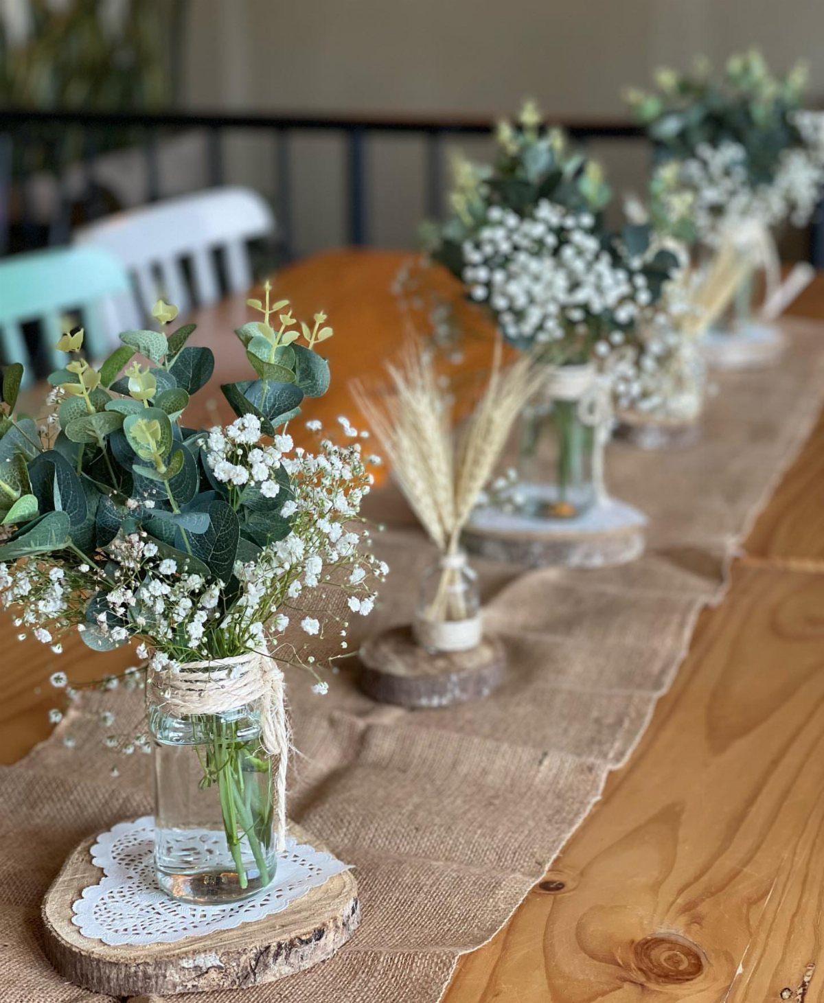 Small glass jars with greenery and baby's breath on wooden slices and burlap.